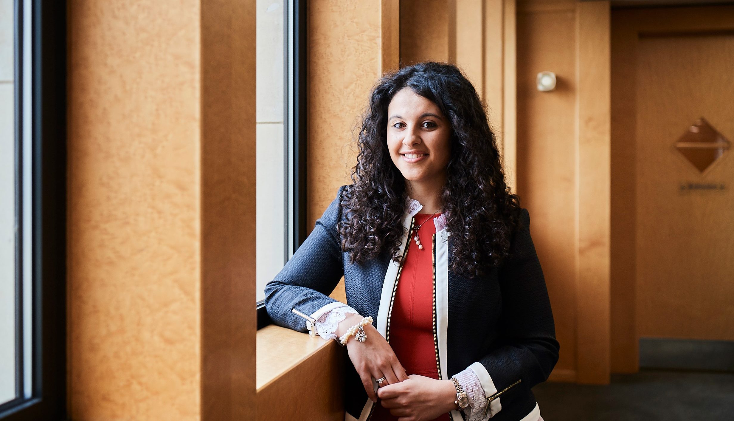 Woman smiling to camera leaning with one elbow on a window ledge in a corridor in an office environment