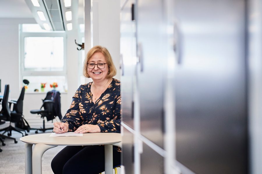Woman smiling at camera whilst sitting at a table close to lockers in office environment